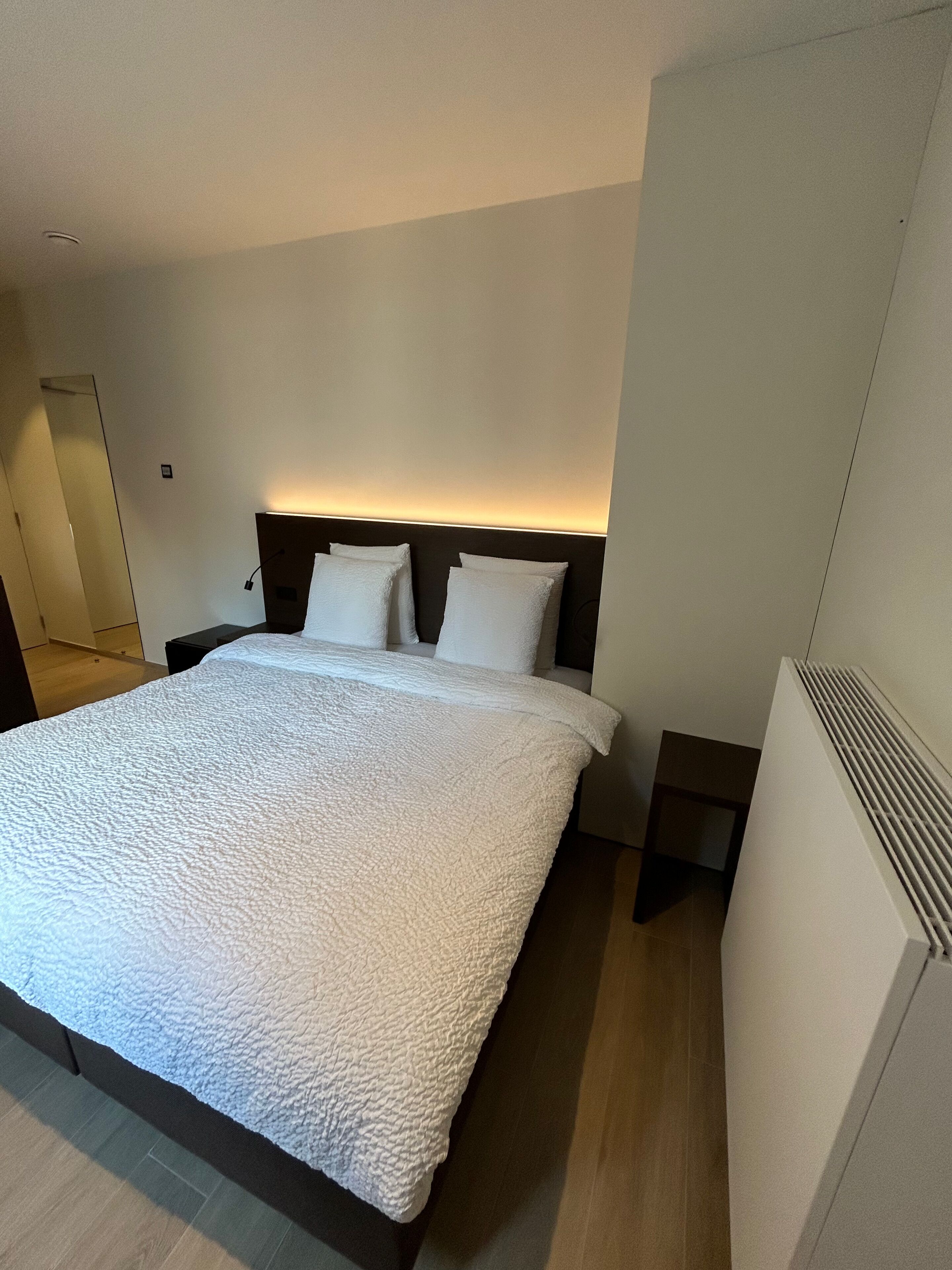 Ein modernes Hotelzimmer mit einem großen Bett, einem mit Beleuchtung versehenen Kopfteil, einem Nachttisch und einer eingebauten Klimaanlage.