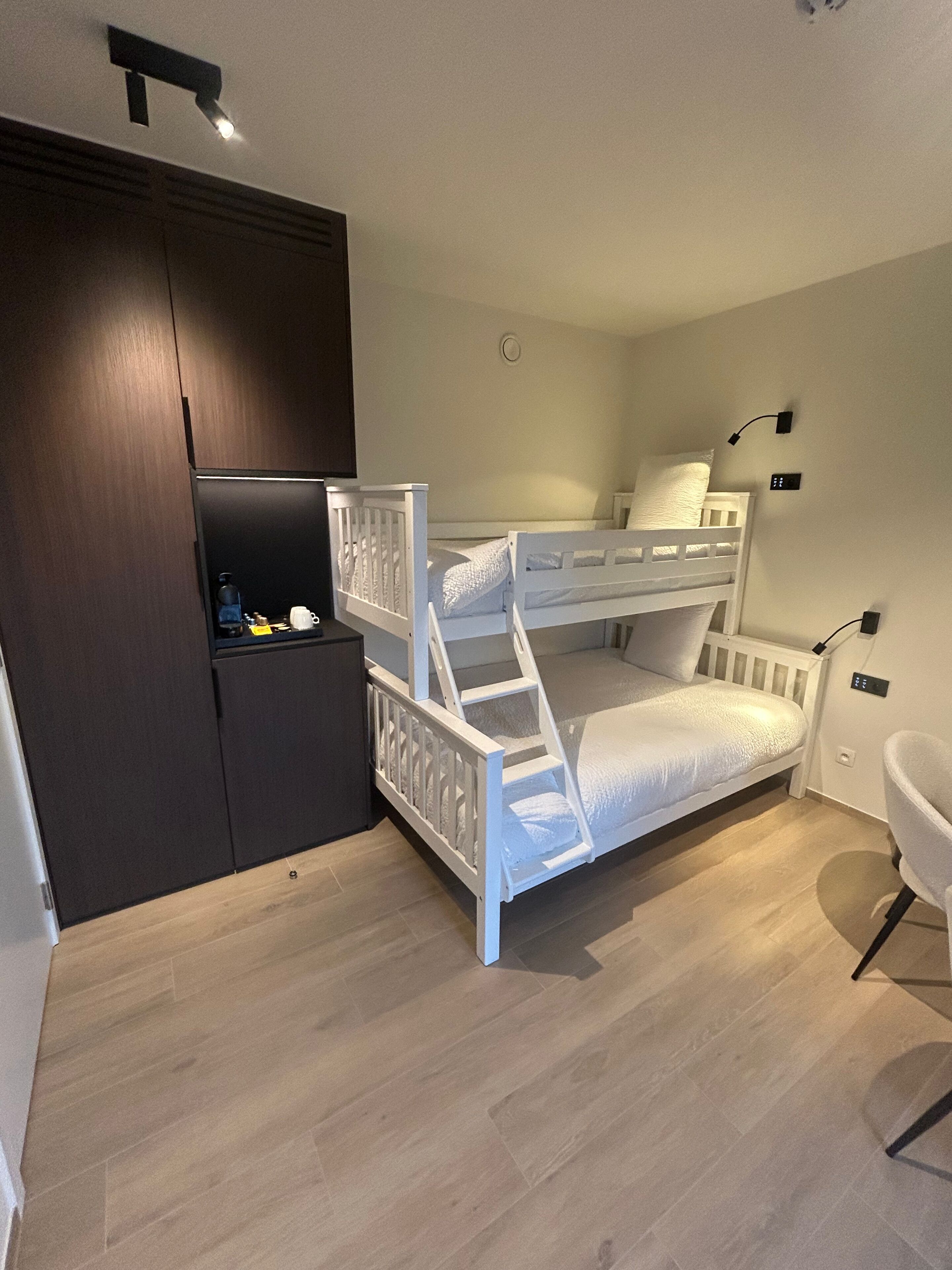 Habitación con literas, un armario oscuro, un estante con objetos y una silla blanca.
