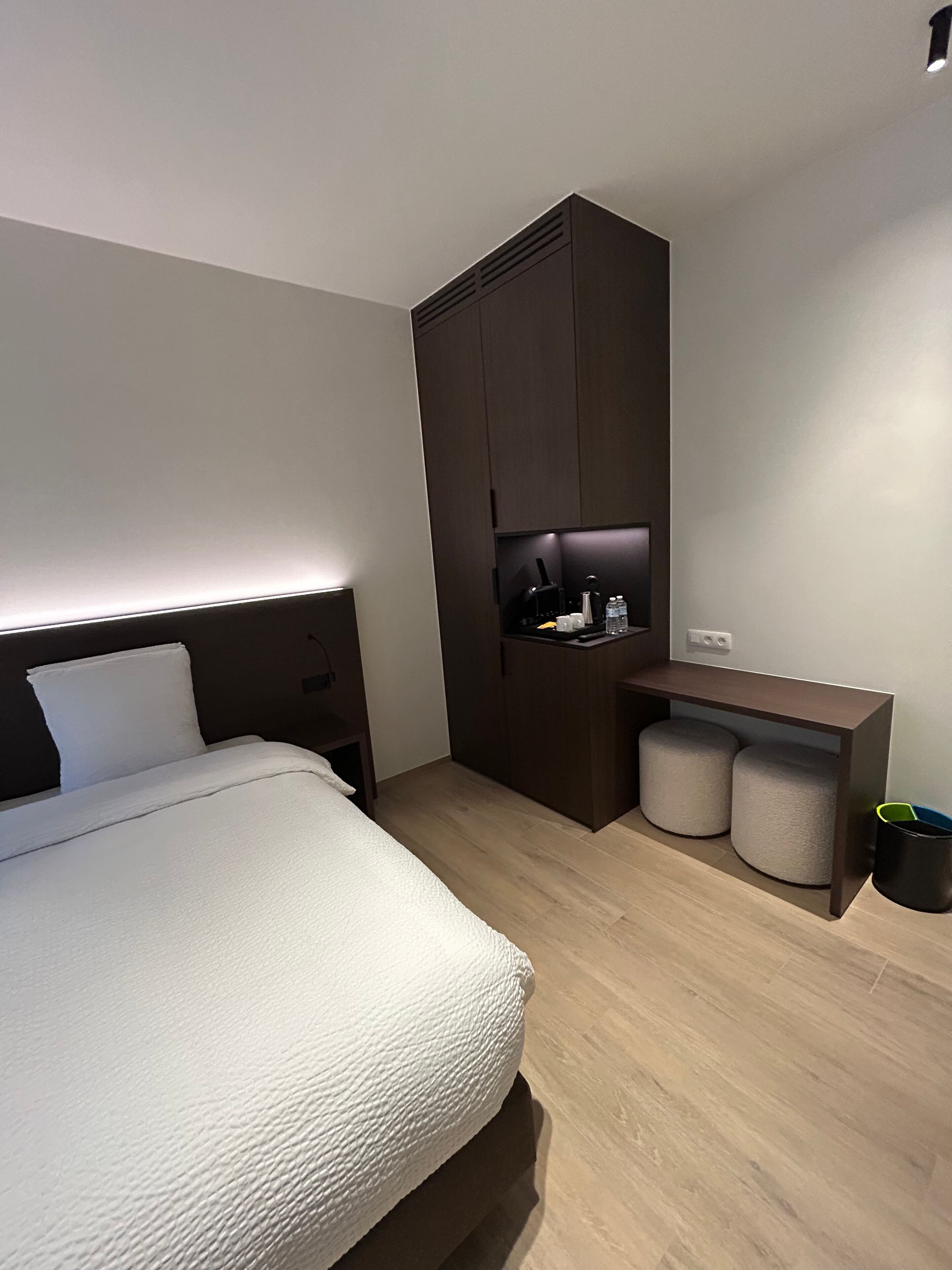 A modern bedroom with a bed, a dark wooden wardrobe, a small desk with a lamp, and a trash bin.