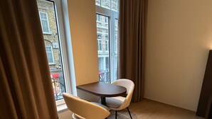 A room with a round table and two chairs, large windows with curtains, and a view of buildings outside. - Manteka Logies (Ypres)