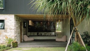 Uma área de estar moderna ao ar livre, com paredes de pedra, um deck de madeira e diversas plantas.