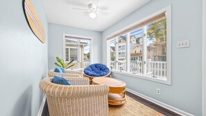 A room with a ceiling fan, wicker furniture, a view of neighboring houses, and a large window.