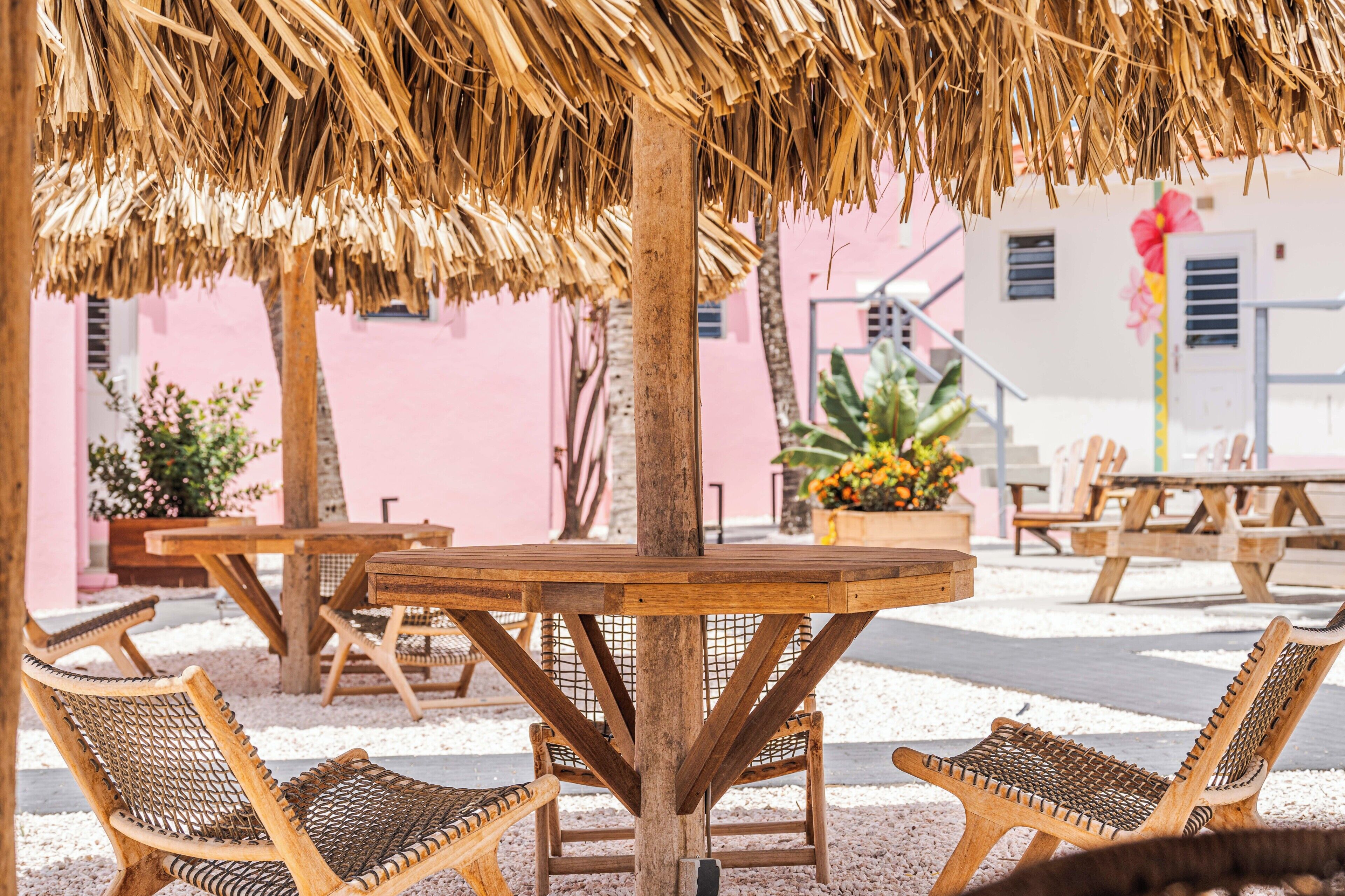 A wooden table and chairs under a thatched roof, with a pink building in the background.