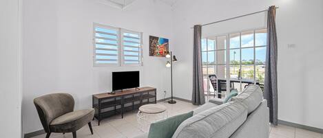 A living room with a sofa, a chair, a TV on a stand, a floor lamp, and a window with curtains.