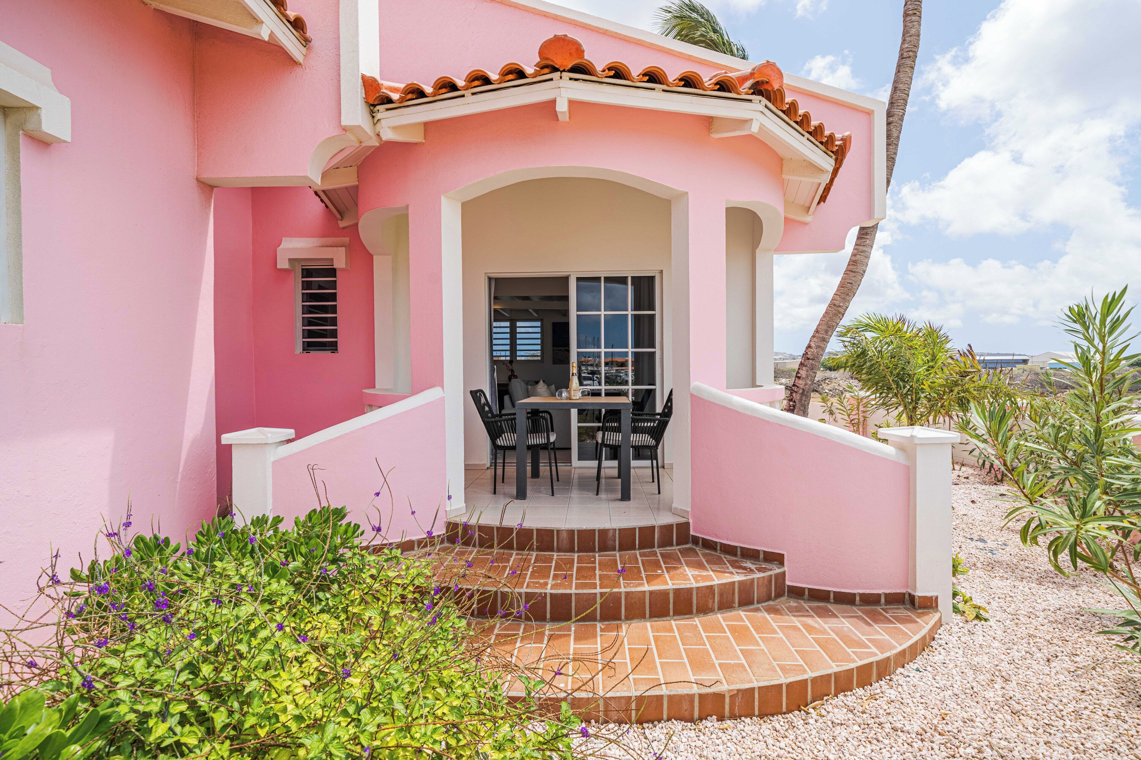 A pink building with a patio area, a table with chairs, and a staircase leading to the entrance.