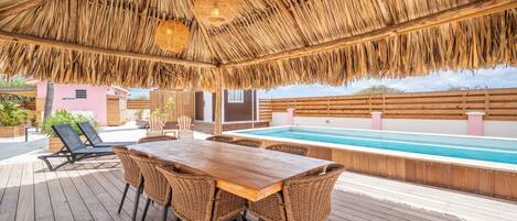 A wooden deck with a thatched roof, a pool, wicker chairs, and a table.