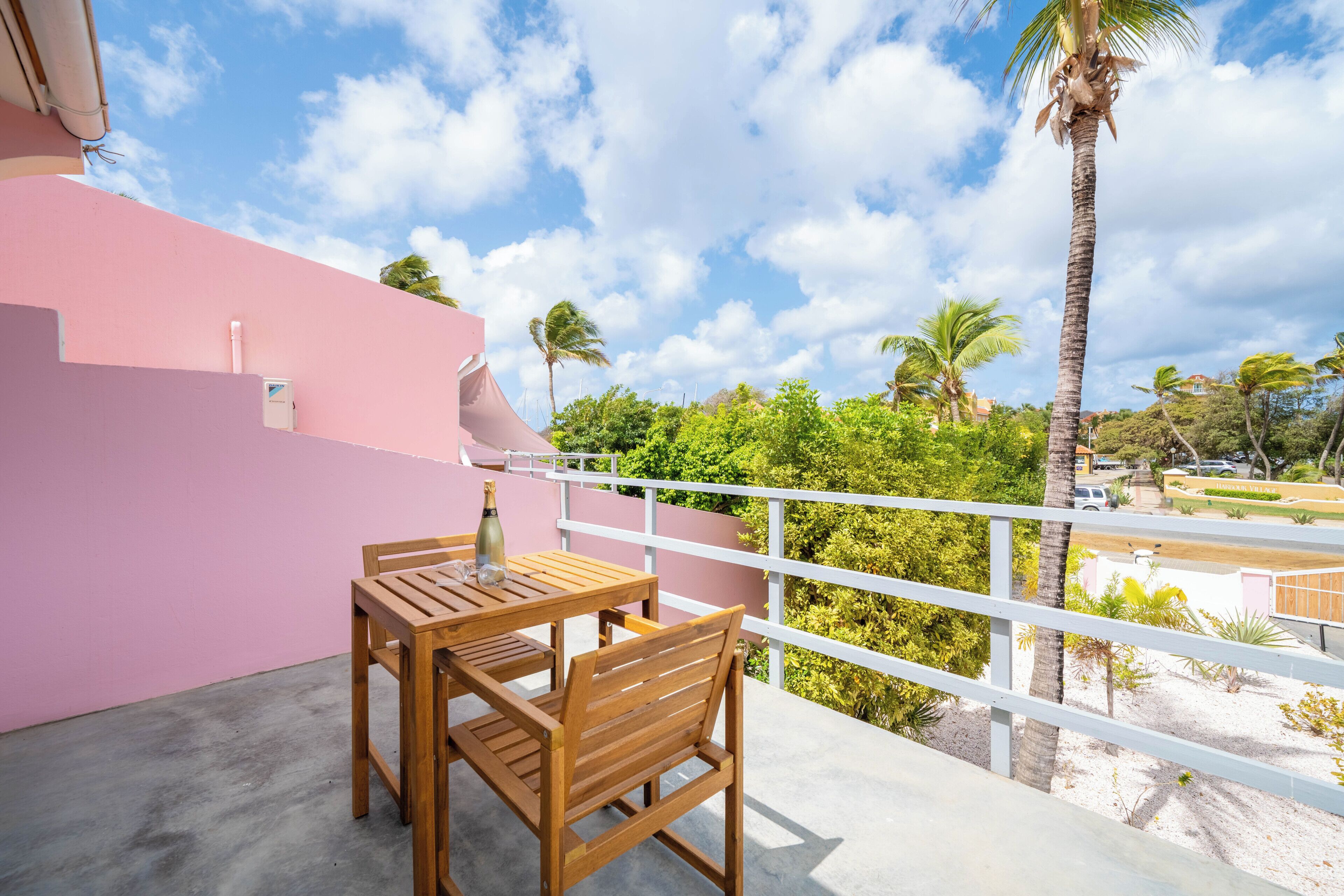 A balcony with a wooden table and chairs, a pink wall, and a view of palm trees and greenery.