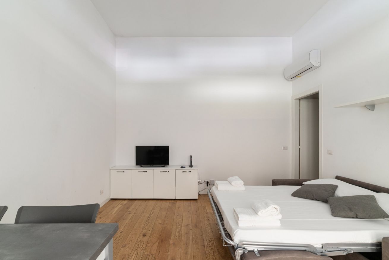 A modern hotel room with a wooden floor, a bed with pillows, a bedside table, a TV, and a wardrobe.