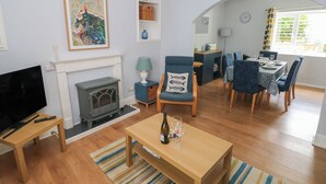 A living room with a fireplace, a television, a dining area with a table and chairs, and a kitchen area with a stove and cabinets. - Longmoor (Tenby)