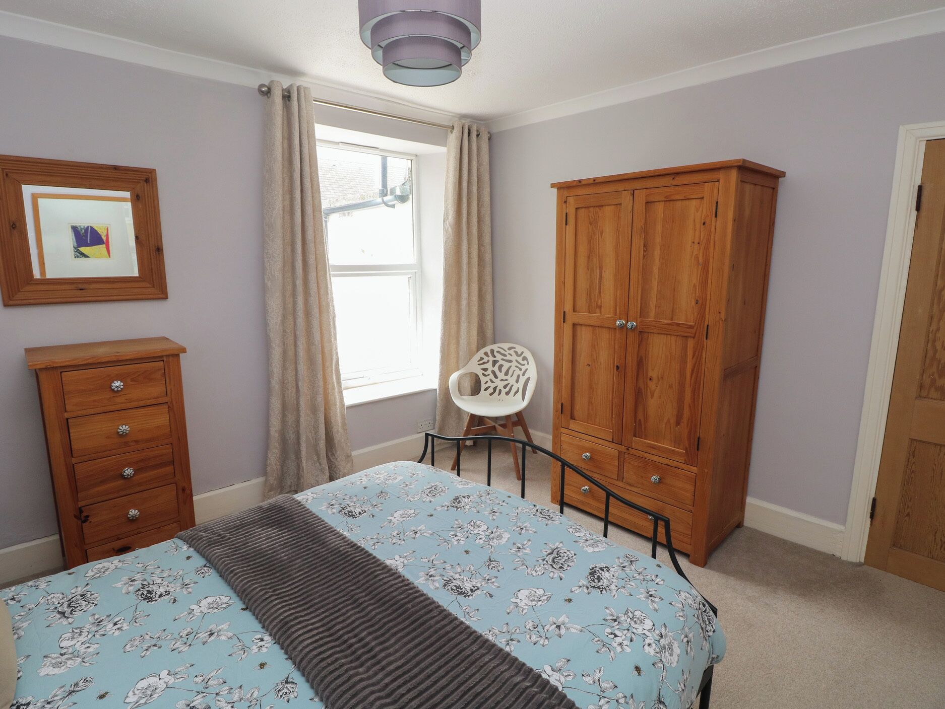 A bedroom with a bed, wooden wardrobe, a chair, a dresser, and a window with curtains.