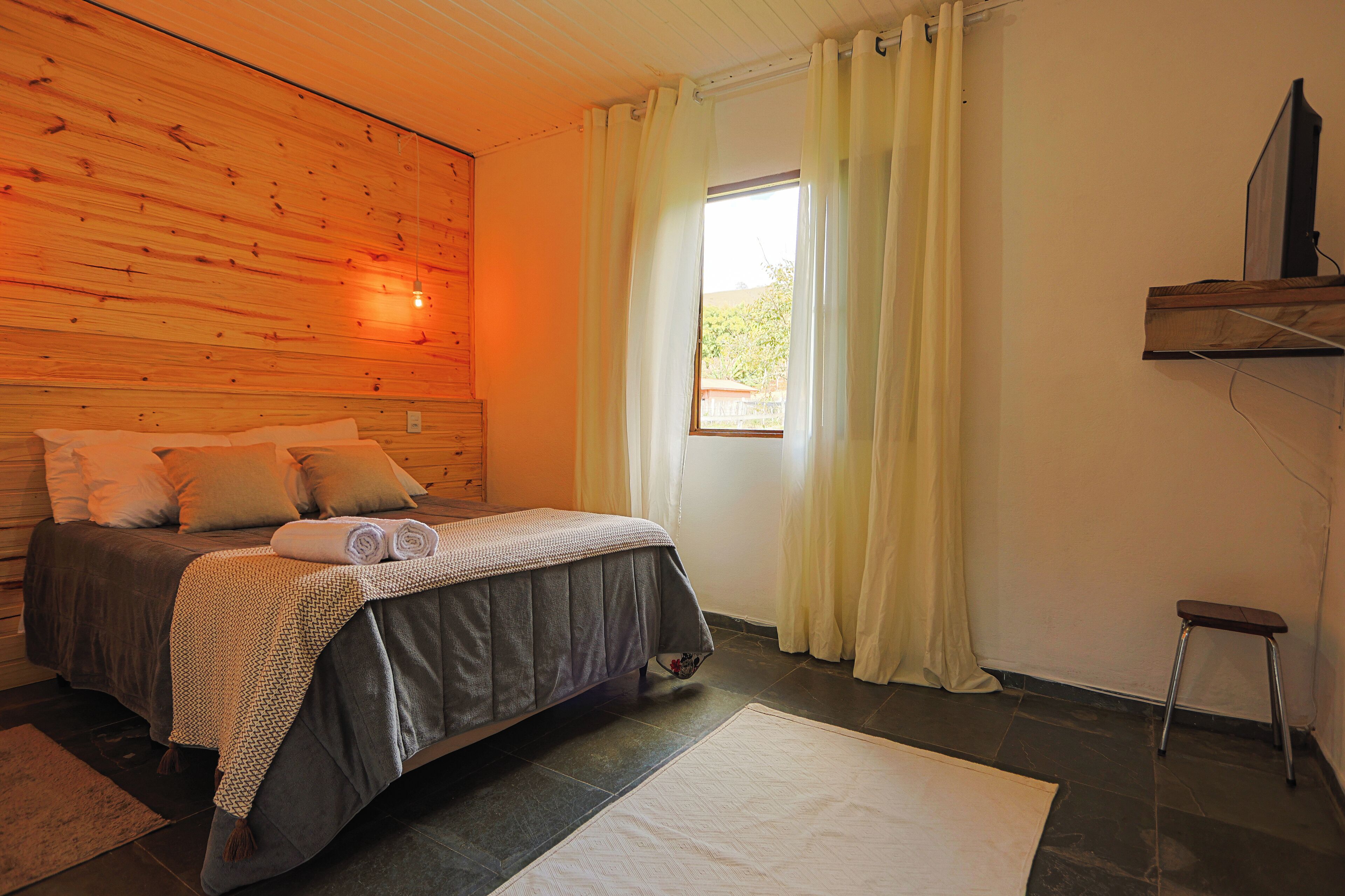 A bedroom with a bed, a small stool, a window with curtains, and a wooden wall.