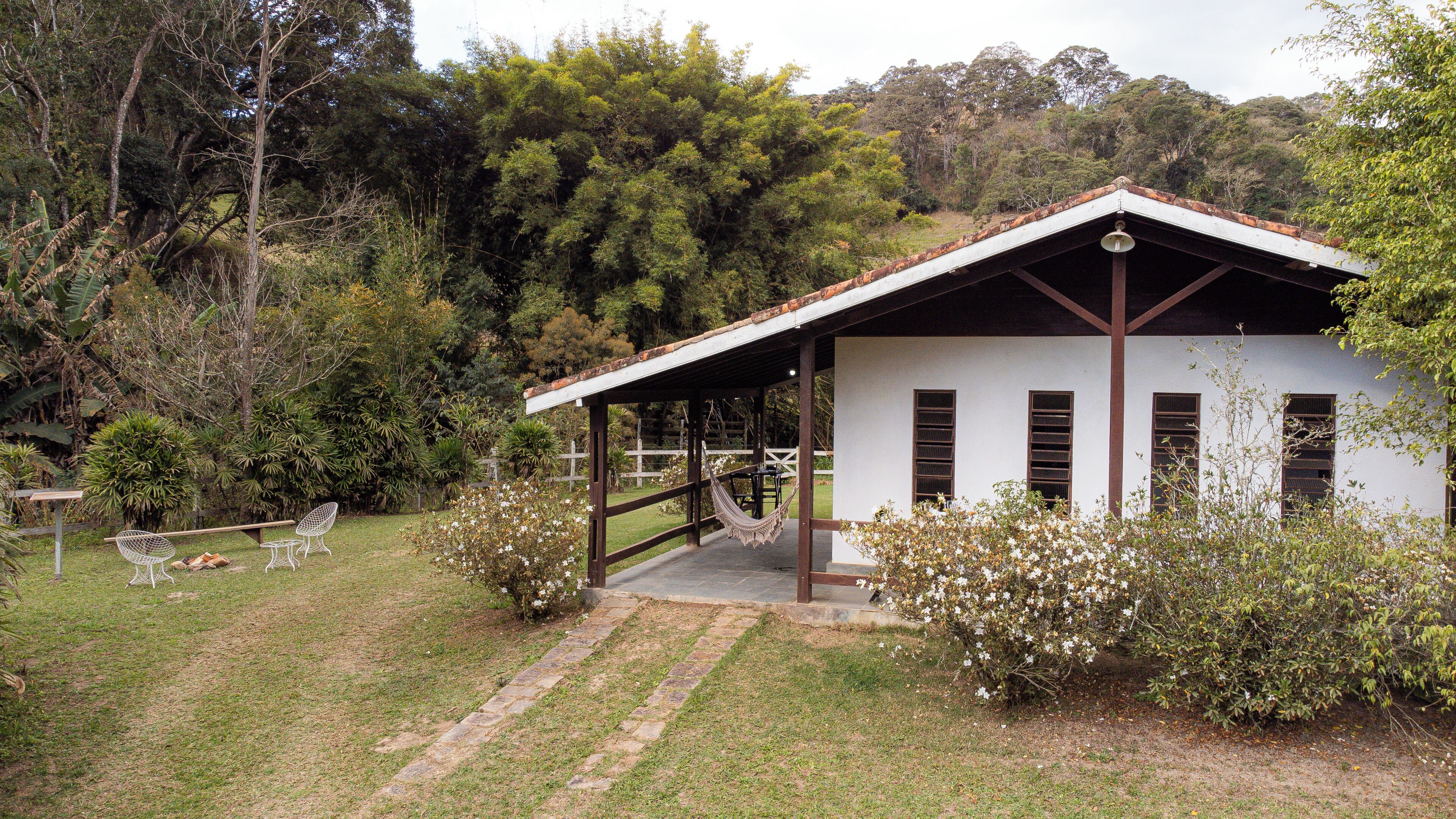 Un petit bâtiment blanc, avec un toit brun et des poutres en bois, entouré d’une végétation luxuriante et d’une zone herbeuse où se trouvent un hamac et des chaises.