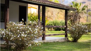 A house with a covered porch, surrounded by lush greenery and hills in the background. - | White cabin | Fireplace, fireplace, tranquility, ideal for families and friends (Gonçalves)
