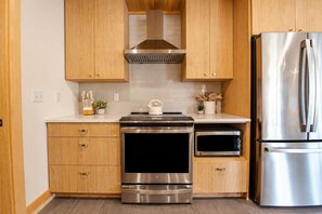 Una cocina moderna con armarios de madera, electrodomésticos de acero inoxidable, incluyendo un refrigerador, horno y microondas, y una placa de inducción negra.