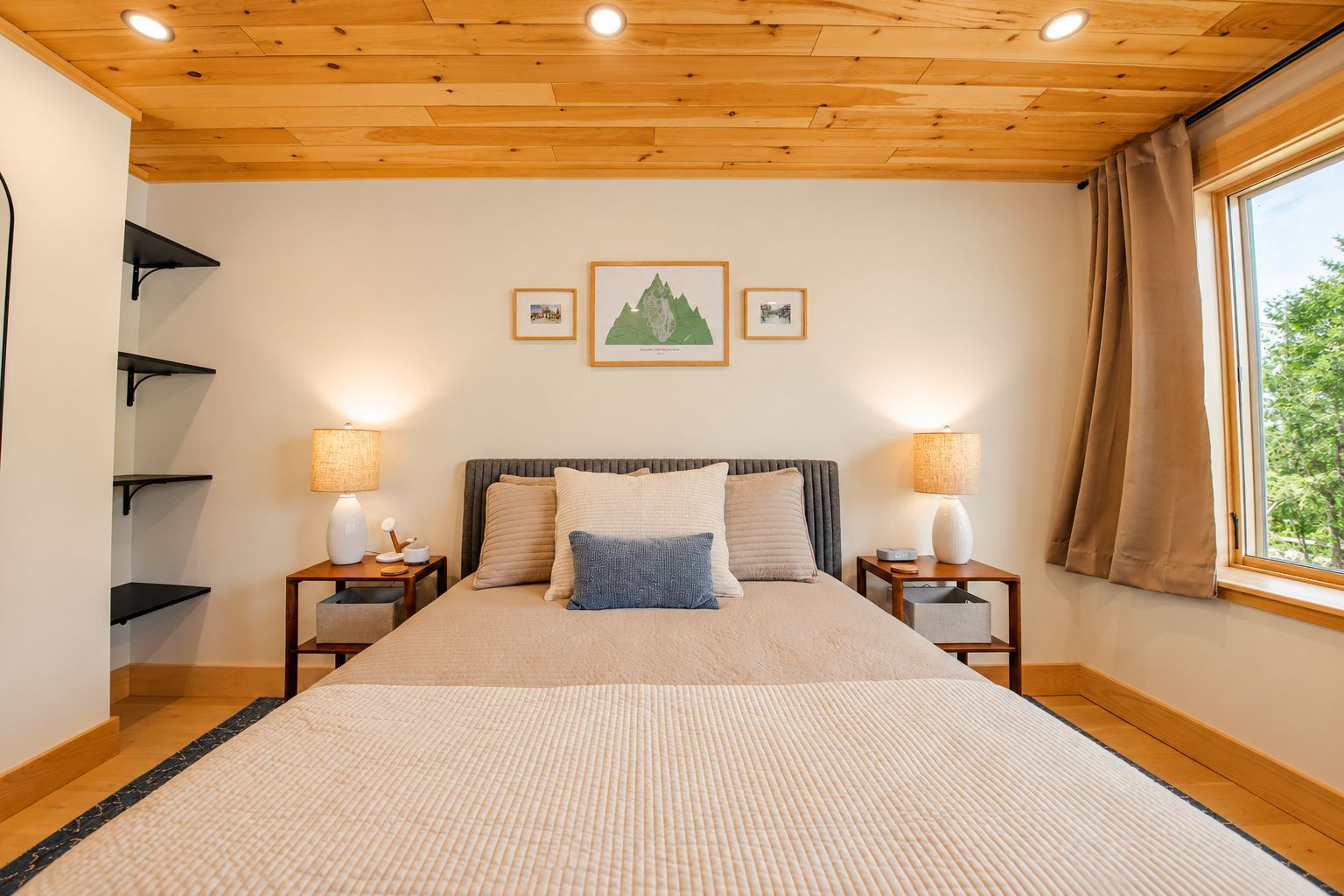 A bedroom with a wooden ceiling, a bed with a beige and blue pillow set, two bedside lamps, and a window with curtains.