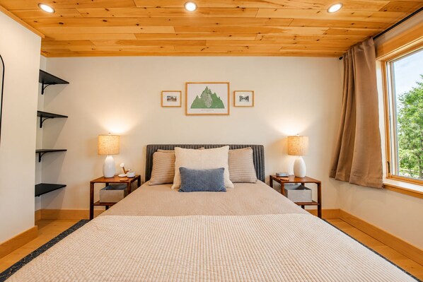 A bedroom with a wooden ceiling, a bed with a beige and blue pillow set, two bedside lamps, and a window with curtains.
