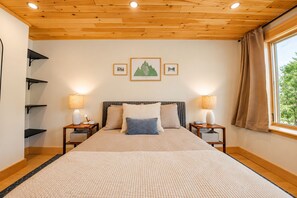 Un dormitorio con techo de madera, una cama con juego de almohadas color beige y azul, dos lámparas de noche y una ventana con cortinas.
