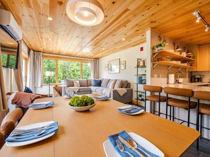 A modern interior with a wooden ceiling, a dining area with a table set for four, a sofa, and a kitchen with bar stools.