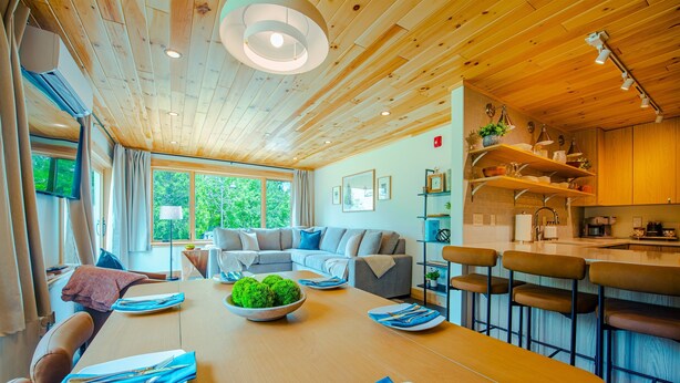A modern, well-lit room with a wooden ceiling, a dining area with a table set for four, a sofa, and a kitchen area with bar stools.