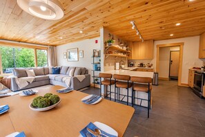 Un interior moderno con techo de madera, una sala de estar con sofá y una mesa de comedor, además de una cocina con taburetes de bar.