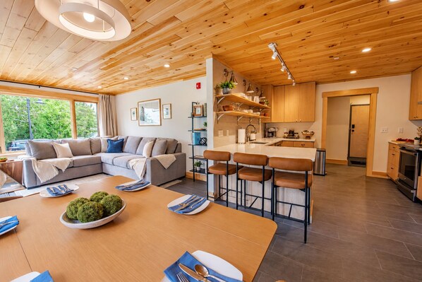 Un interior moderno con techo de madera, una sala de estar con sofá y una mesa de comedor, además de una cocina con taburetes de bar.