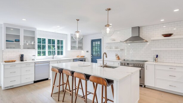 Une cuisine moderne avec des meubles blancs, un îlot central doté de tabourets, une cuisinière et des luminaires suspendus.