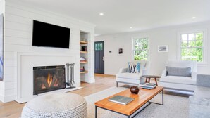 Un salon moderne doté d’une cheminée, d’un téléviseur à écran plat fixé au-dessus, et d’un coin salon confortable avec un canapé et des fauteuils.