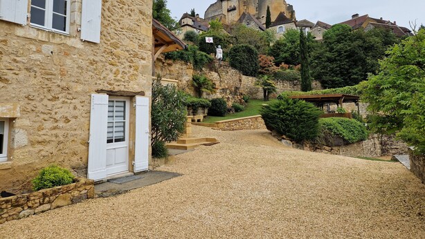 A stone building with a gravel driveway leading to a castle-like structure in the background. - CHAMBRE D'HÖTE AT THE FOOT OF THE CHATEAU SLIDING-SCALE RATES PLUS 2 NIGHTS WEEKEND INCLUDED (Castelnaud-la-Chapelle)