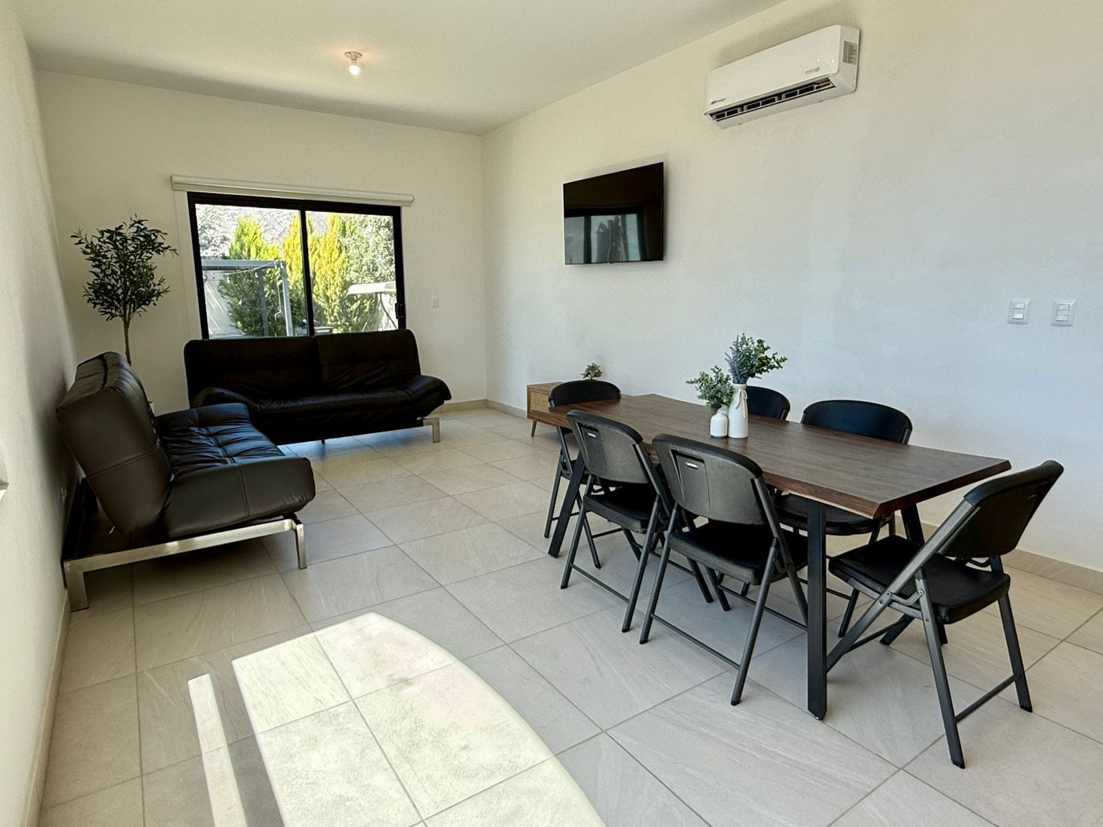 Una sala de estar moderna con un sofá, una mesa de comedor con sillas, un televisor montado en la pared y un equipo de aire acondicionado.