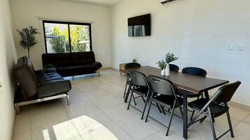 Una sala de estar moderna con un sofá, una mesa de comedor con sillas, un televisor montado en la pared y un equipo de aire acondicionado.