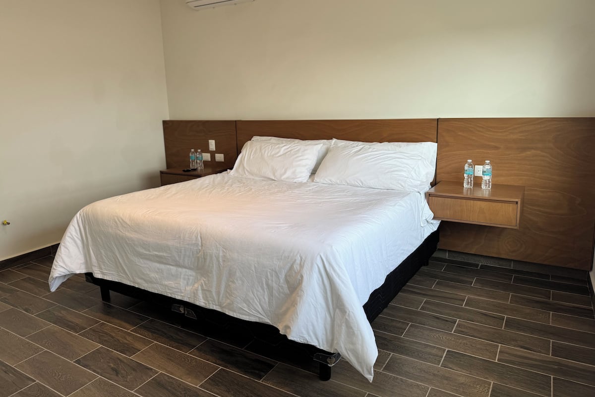 Een net opgemaakt bed met witte lakens, een houten hoofdbord en een nachtkastje met twee waterflessen.