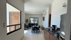 A modern kitchen with a stove, sink, and cabinets, a dining area with a table and chairs, and a living room with a sofa and a plant. - Departamentos Aremay (Cuatro Ciénegas)