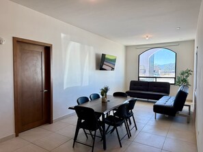 A modern living room with a dining area, a sofa, a TV mounted on the wall, and a large window offering a view.