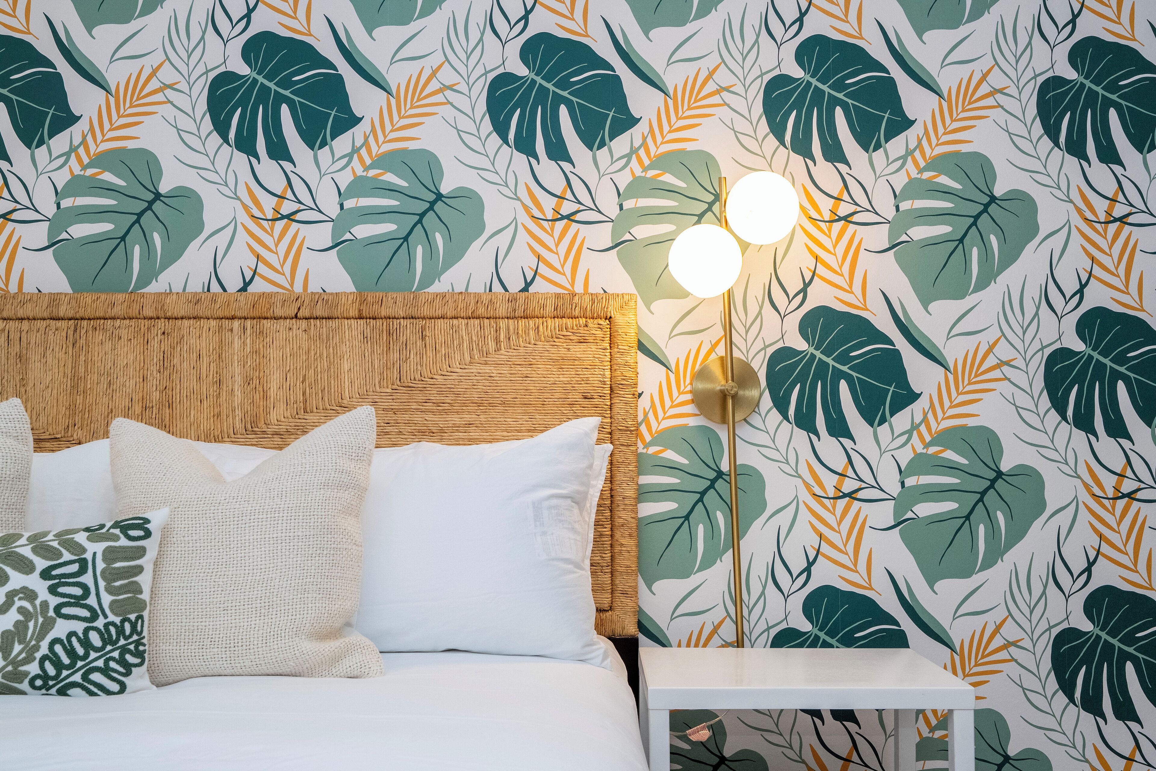 A bed with white bedding and multiple pillows, a wooden headboard, a nightstand with a lamp, and a wall with a tropical leaf pattern.