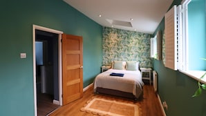 Ein Schlafzimmer mit türkisfarbenen Wänden, einer Holztür, einem Bett mit weißer Bettdecke, einem Nachttisch, einem kleinen Tisch und einem Fenster mit Jalousien.
