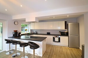 A modern kitchen with a central island, bar stools, stainless steel appliances, and a clock on the wall. - Spacious 5-Bedroom Home with Large Garden in Egham (Egham)