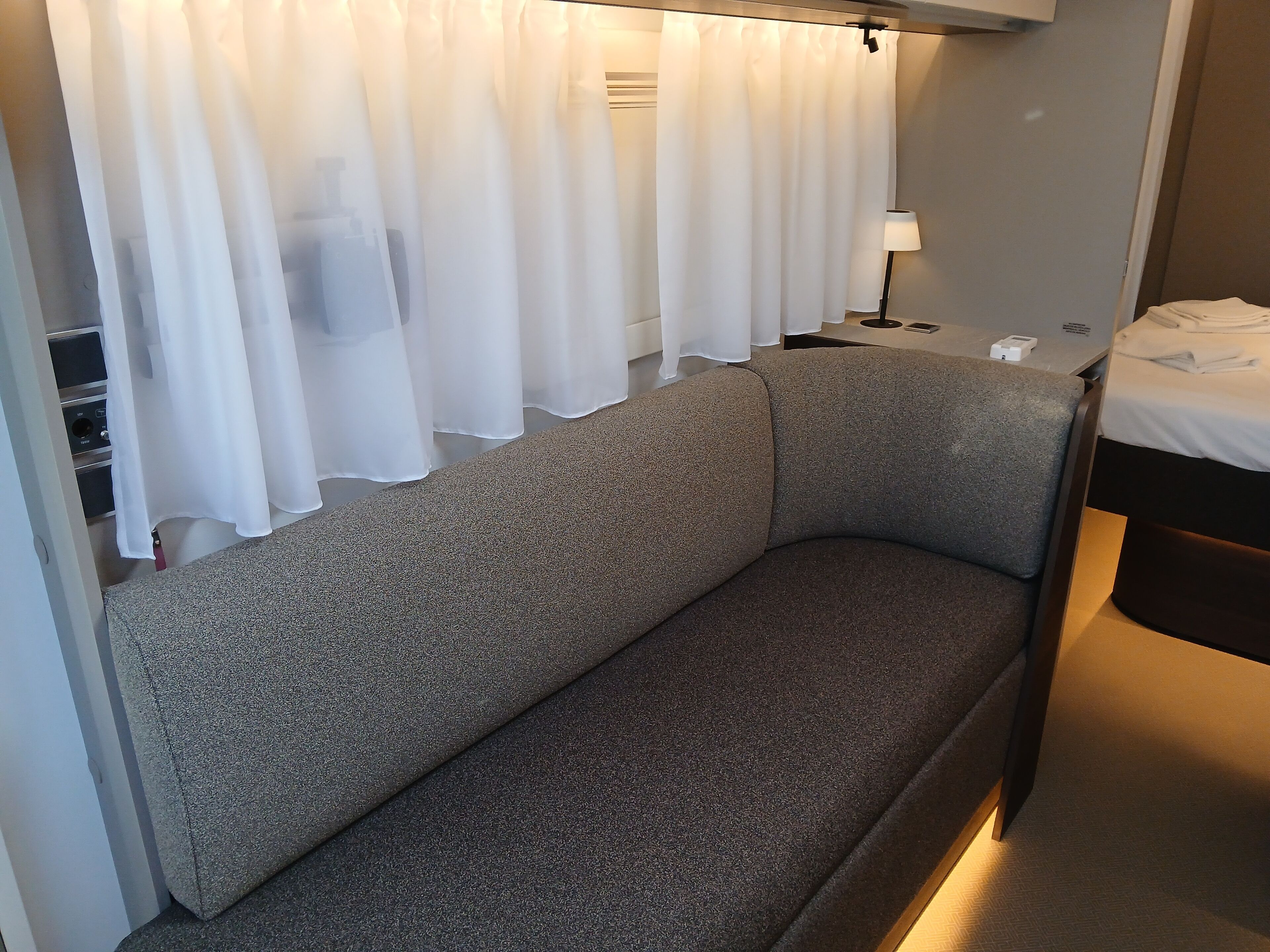 Habitació moderna i compacte amb un sofà gris, una taula de nit petita amb una làmpada, i un llit fet amb cura.