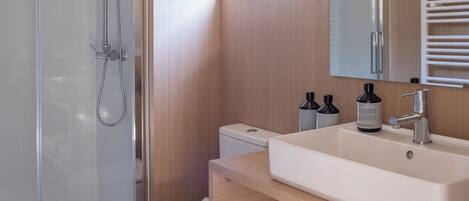 Un bagno moderno con doccia vetro, vaso bianco, un ampio lavandino con specchio sopra e asciugamani piegati ordinatamente su uno scaffale.
