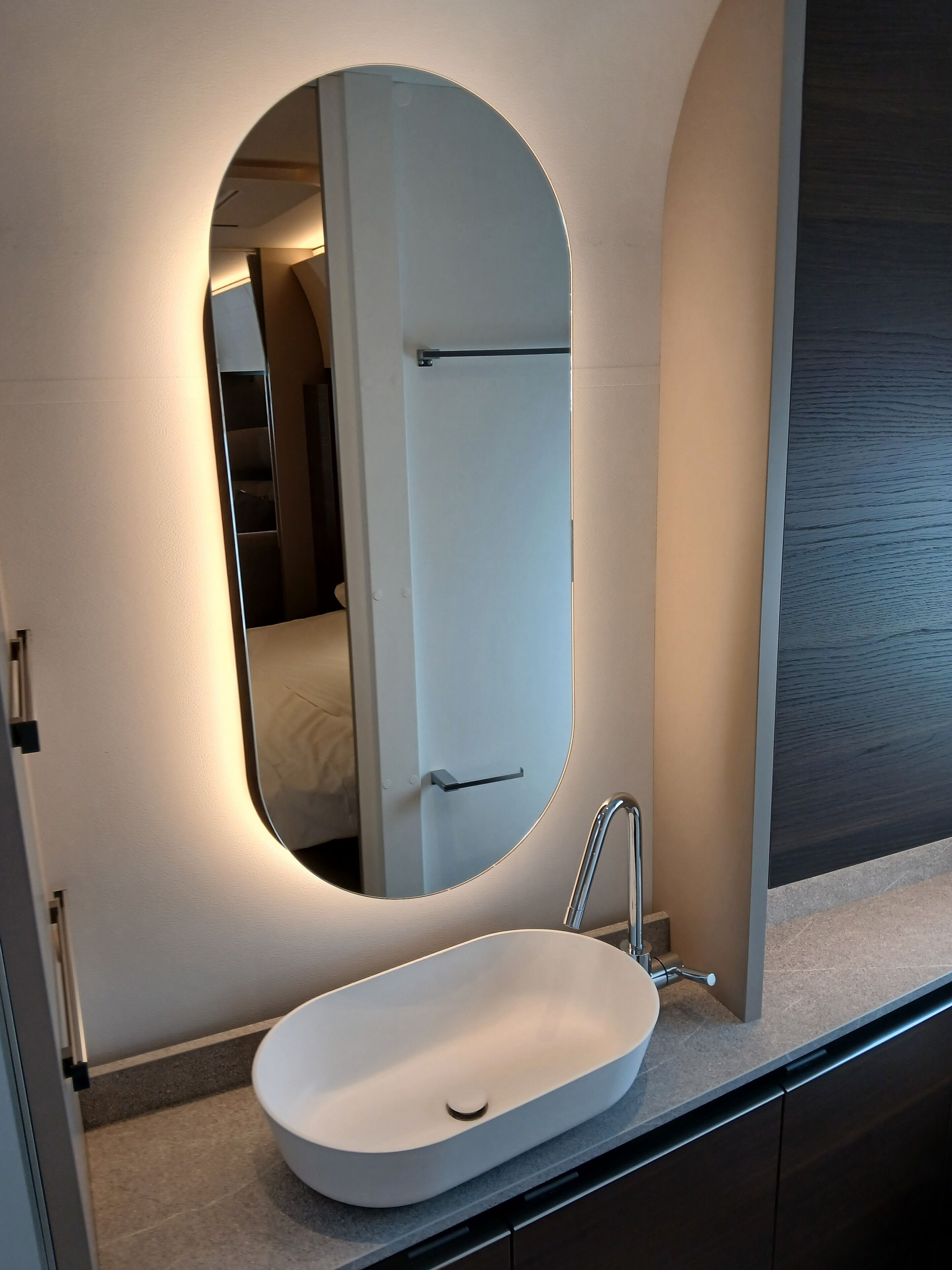 Une salle de bain moderne avec un lavabo ovale blanc, un grand miroir et une porte donnant sur une chambre.