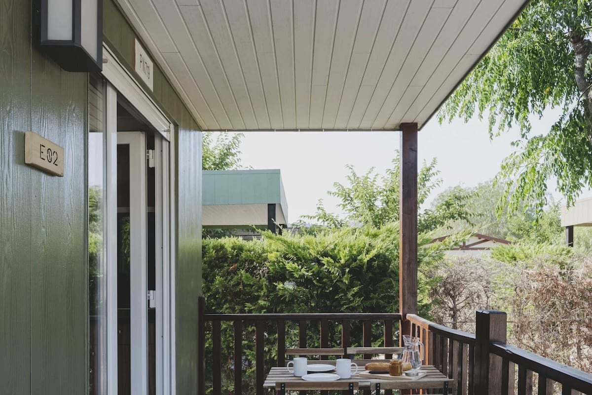 A wooden deck with a table set for dining, a sign reading "E02", and a view of a building and greenery in the background.