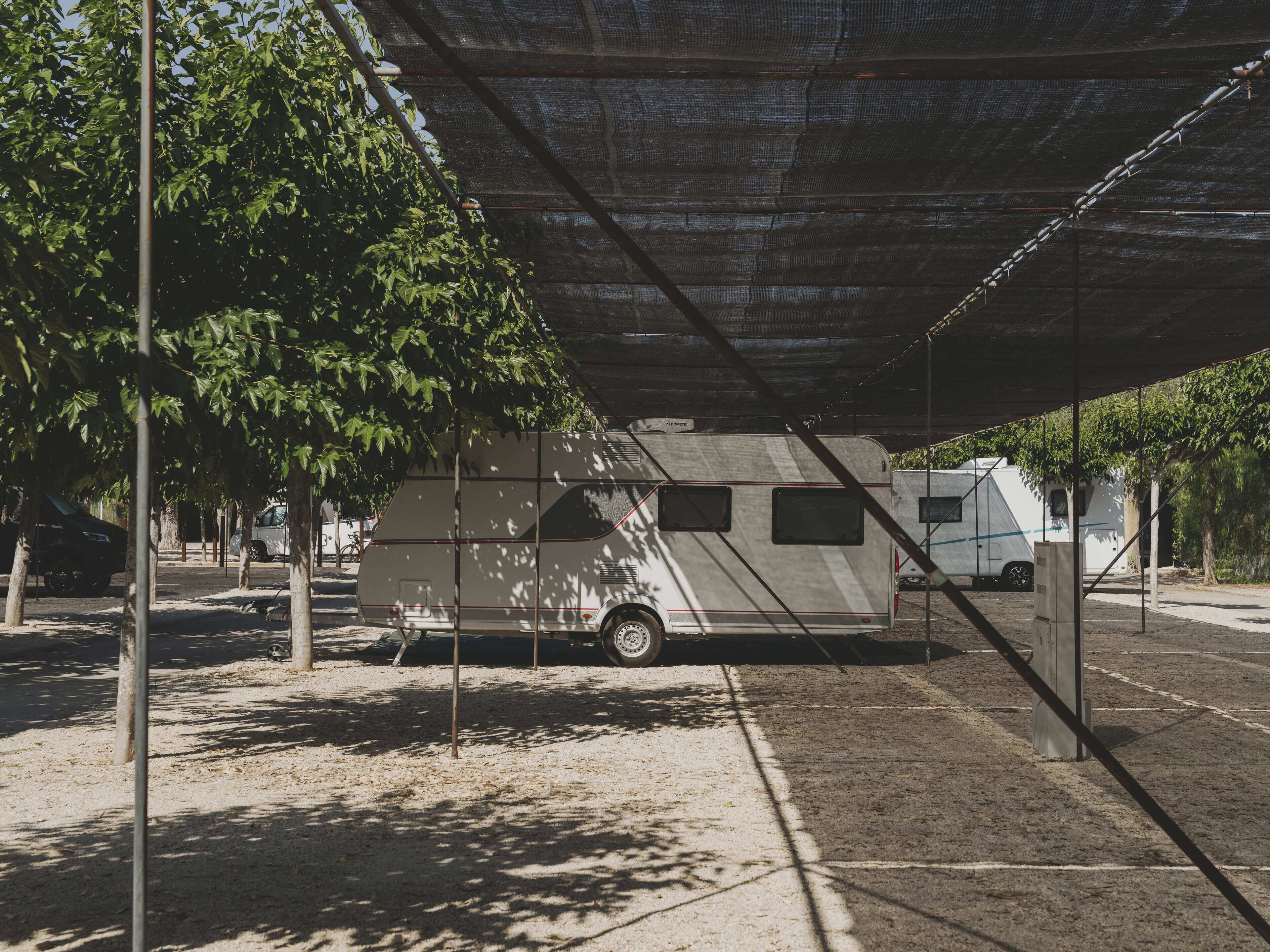 Área de estacionamento coberta para veículos recreativos, com árvores e chão de terra.