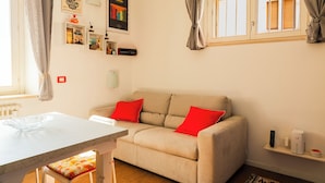 A cozy living room with a beige sofa, red cushions, a small table, and a window with curtains.