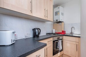 A kitchen with light wooden cabinets, a black countertop, a white toaster, a black kettle, a red appliance, and a stainless steel oven.
