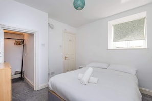 A bedroom with a bed, a window with a floral blind, a closet with hanging clothes, and a door.