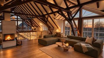 Een ruime loft met zichtbare houten balken, een open haard, een grote hoekbank en uitzicht door de grote ramen.