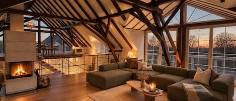 Un loft espacioso con vigas de madera a la vista, una chimenea, un amplio sofá modular y vistas a través de amplios ventanales.