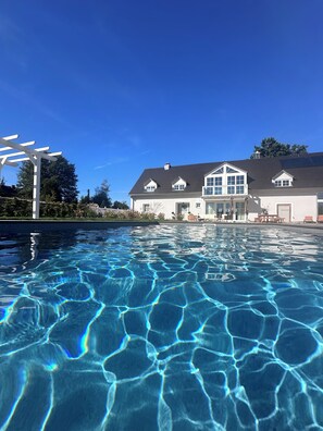 Ein Schwimmbad mit klarem Wasser, das den Himmel spiegelt, eine weiße Pergola und ein großes Haus mit dunklem Dach und mehreren Fenstern.