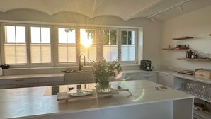Et moderne køkken med en stor ø, hvide skabe og udsigt til naturen gennem store vinduer.