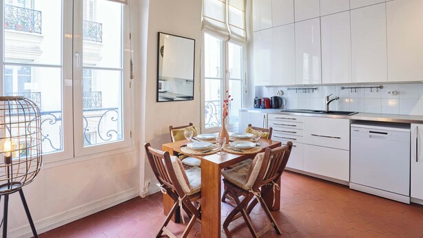 A dining area with a wooden table set for four, white cabinets, a kitchen with a sink and dishwasher, and large windows with blinds. - Charming Apartment  with AC- Heart of Le Marais (Paris)