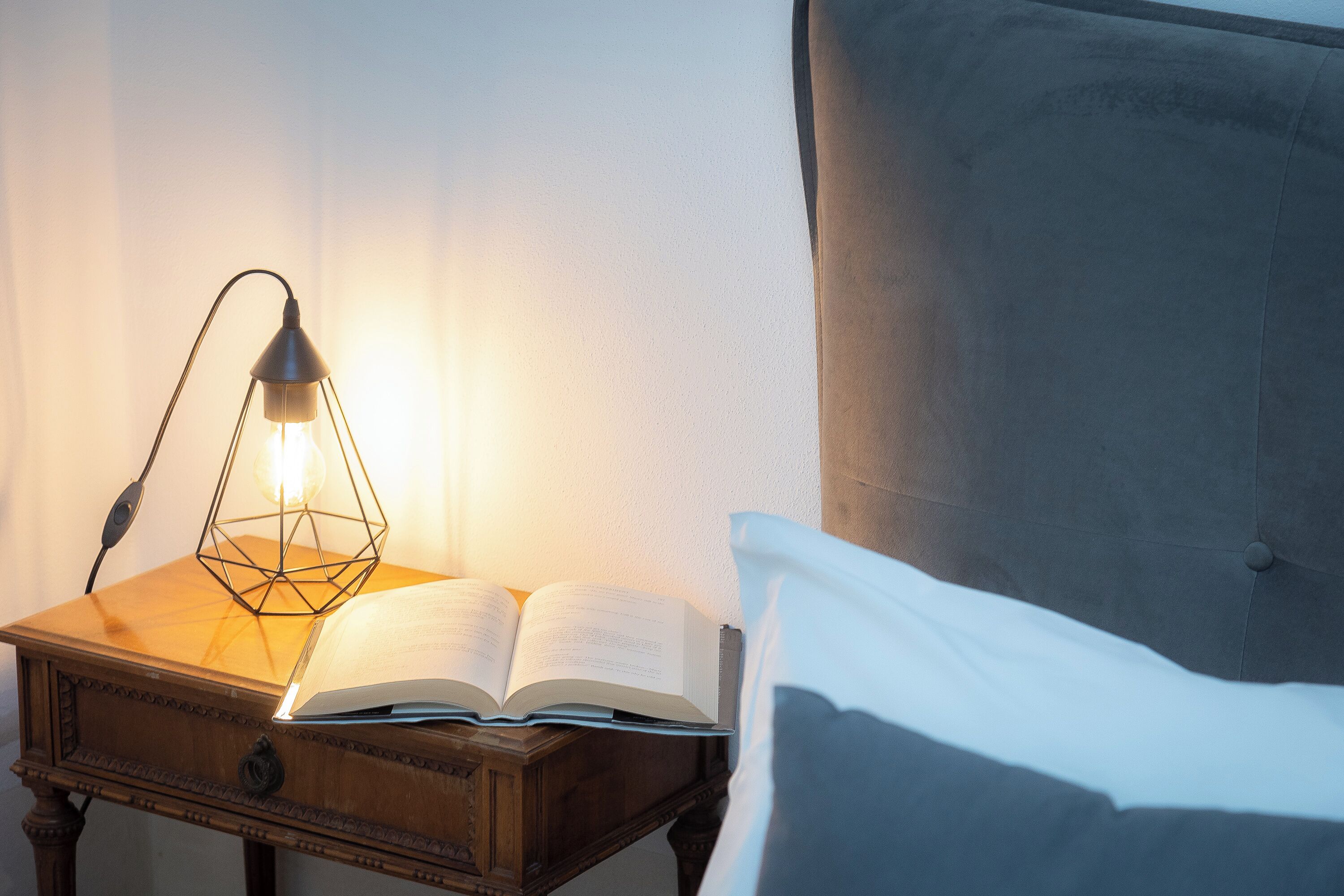 A bedside table with an open book and a lamp, next to a bed with a pillow.